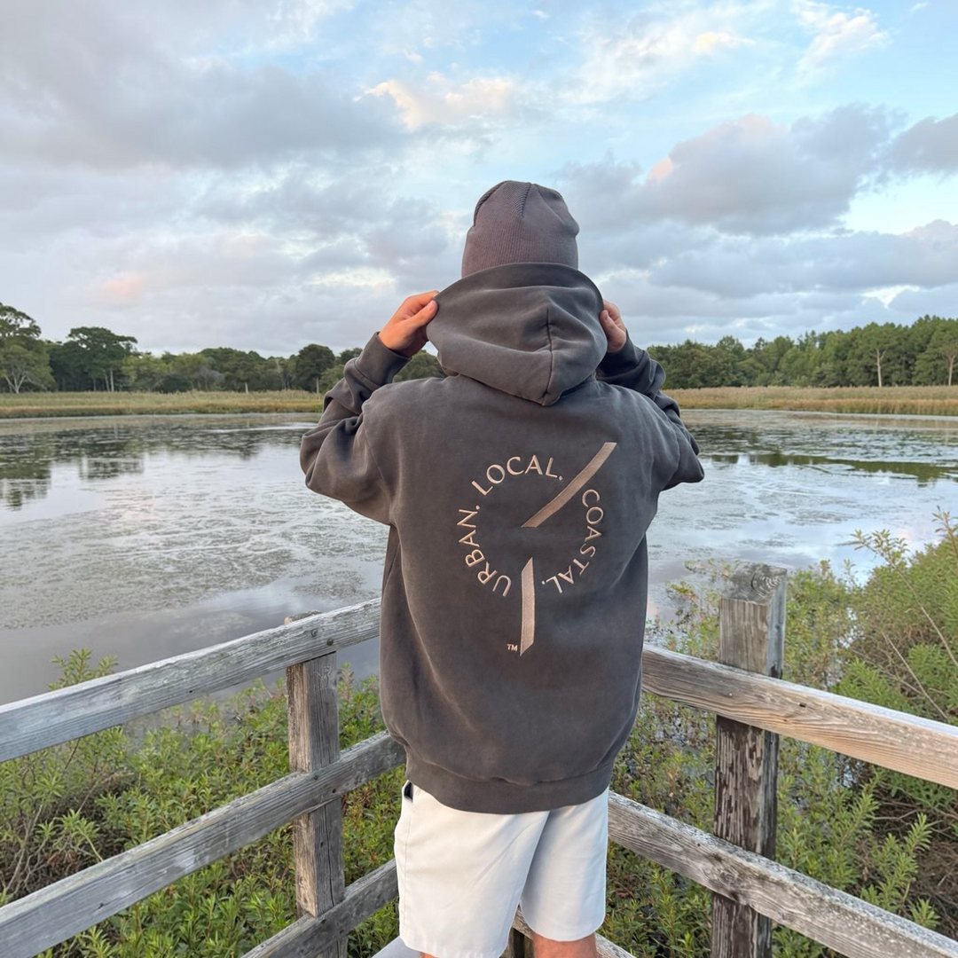 area 609 The Founders Hoodie in pigment black with back embroidery, worn by a person in a beanie by a lake