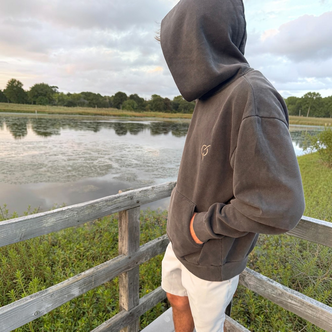 area 609 heavyweight pigment black hoodie with core emblem on chest, worn by a person on a wooden pier by a lake